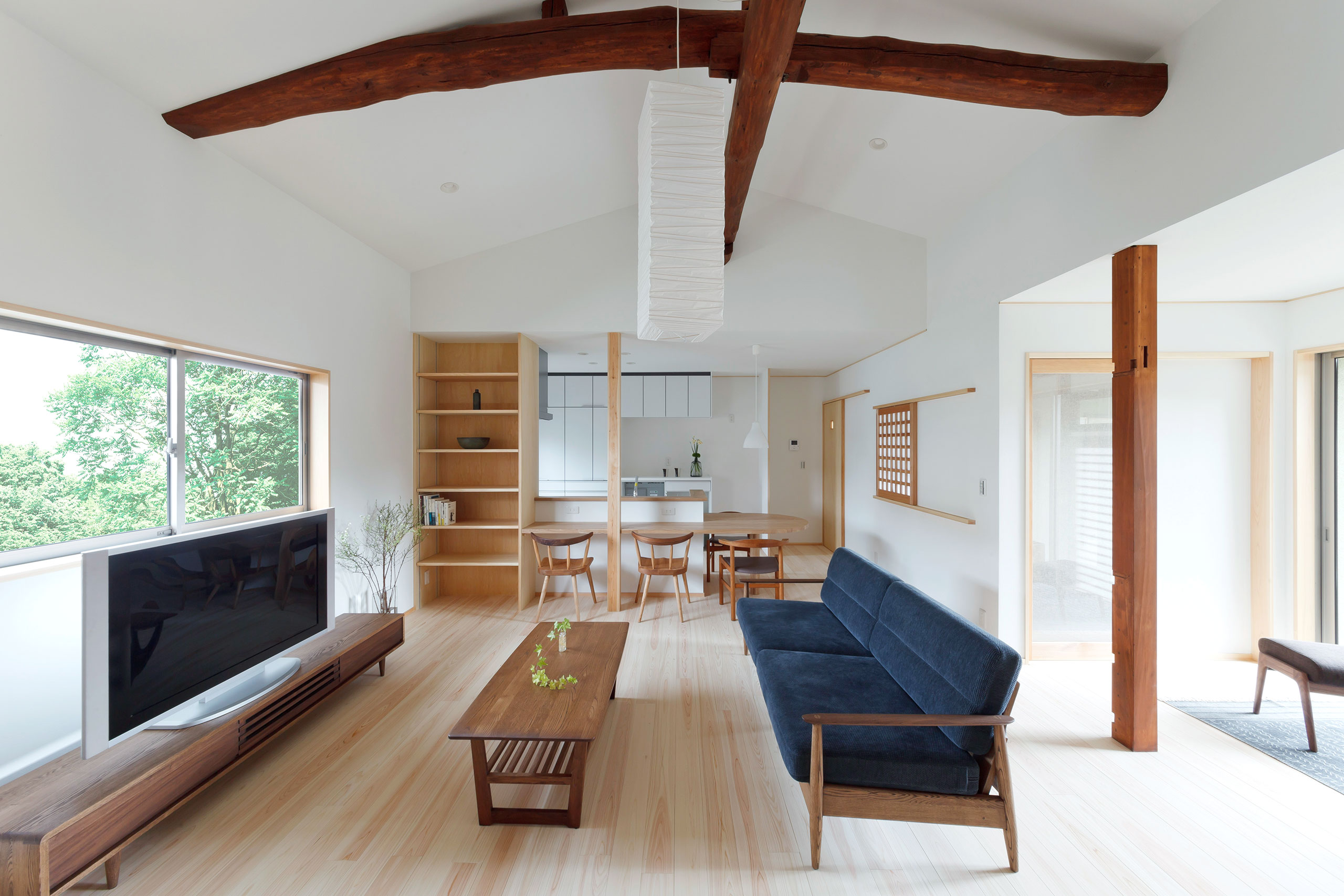 木製家具とネイビーのソファが配置された、勾配天井と大きな窓のあるナチュラルなリビングダイニング空間